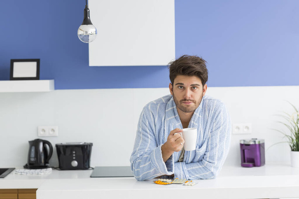 Portrait of young ill man holding coffee mug while leaning on kitchen counter - Photo, image