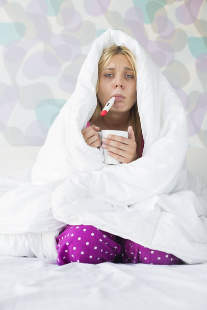 Portrait of young woman with coffee mug taking temperature while wrapped in quilt on bed - Fotó, kép