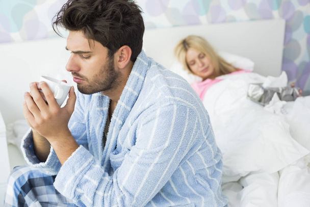 Sick man drinking coffee on bed while woman sleeping in background at home - Fotoğraf, Görsel