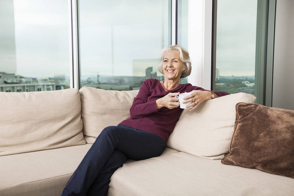 Smiling senior woman with coffee mug relaxing on sofa at home - Foto, Imagen