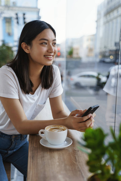 Vertical outdoor shot cheerful beautiful young modern asian girl in t-shirt, jeans, lean coffee table, holding smartphone, dreamy contemplate city streets through window, messaging waiting for friend - Foto, Imagen