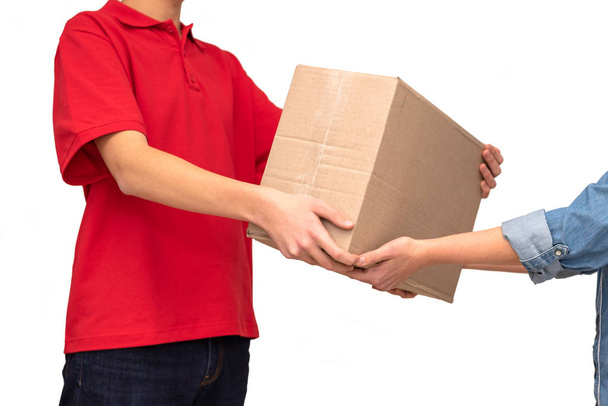 Vrouwelijke handen ontvangen pakketje van bezorger geïsoleerd op witte achtergrond. Jonge koerier gekleed in rood T-shirt geeft kartonnen doos. Bezorgingsconcept. Kopieerruimte. - Foto, afbeelding