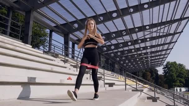 Attractive slender sporty young woman with ponytail in black fitness clothes doing stretching exercises with elastic special band outdoors - Πλάνα, βίντεο