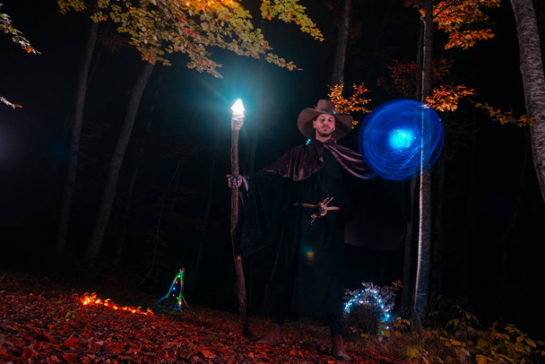 Čaroděj (kouzelník) seslal kouzlo, když stál v kouzelném lese. Držel svou kouzelnou hůl. Magické osvětlení. Podzimní svátky. Mystery and nightmare concept - Fotografie, Obrázek