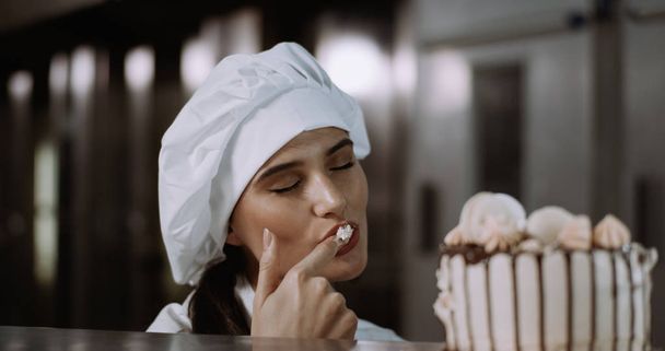 Smiling excited young baker woman in a industrial kitchen bakery she tasting the cream from the fresh baked cake - Фото, изображение