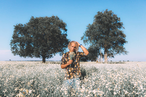 Ritratto di ragazzo biondo riccio è in piedi sul campo e annusa un fiore. Romantico, periodo estivo. Fiori intorno e alberi
. - Foto, immagini