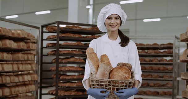 Hermosa señora panadero en un uniforme blanco sosteniendo una cesta con pan fresco y sonriendo lindo delante de la cámara
 - Foto, Imagen