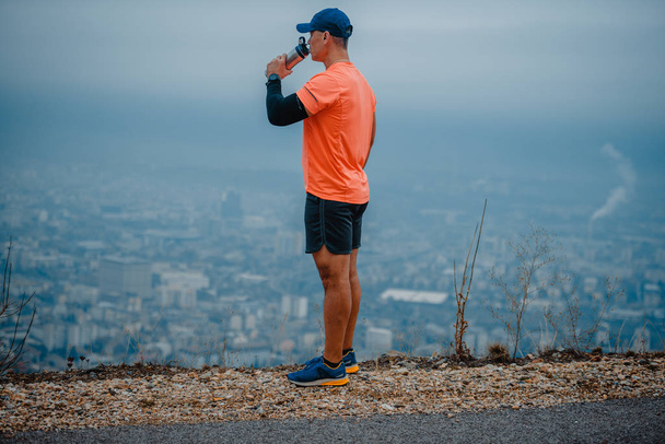 Portrait de mâle sportif mature en vêtements de sport colorés se reposant après l'entraînement de remise en forme, boire de l'eau de la bouteille sur fond bleu ciel
 - Photo, image