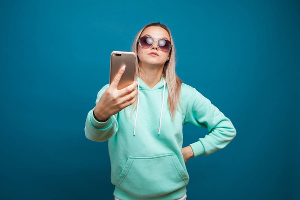 Cheerful blonde with a mobile phone. Portrait of a young positive woman in a blue hoodie - Fotoğraf, Görsel