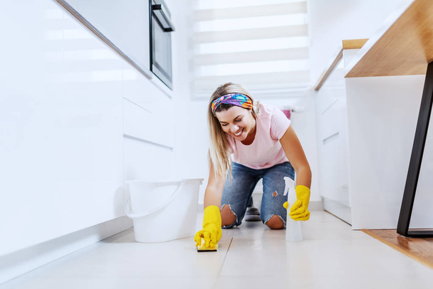 Dedicated smiling beautiful caucasian blond housewife with rubber gloves on kneeling and cleaning kitchen floor with detergent and sponge. Next to her is bucket with water. - Photo, Image