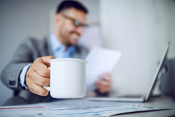 Beautiful caucasian classy smiling businessman in suit and with eyeglasses holding in one hand coffee and in other document and looking at it. Selective focus on mug with coffee. Office interior. - Foto, Imagem