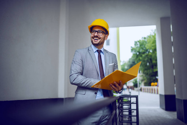 Úspěšný staveniště manažer stojí venku a drží žlutou složku v šedém obleku a s ochrannou přilbou na hlavě - Fotografie, Obrázek