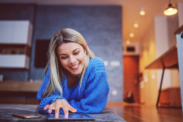 Beautiful smiling Caucasian blond woman in sweater lying on stomach on living room floor with bluetooth earphones and using tablet. - Photo, Image