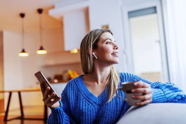 Charmante blanke blonde vrouw zittend in de woonkamer op de bank, met een kopje koffie en een smartphone en kijkend door het raam. - Foto, afbeelding