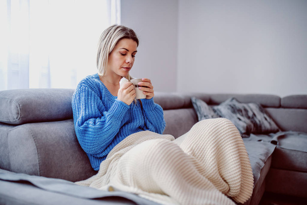 Ill cute caucasian blond woman covered with blanket sitting on sofa in living room, drinking tea and holding handkerchief. - Photo, Image