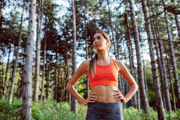 Fit slim sporty serious caucasian brunette in sportswear and with ponytail standing in the woods with hands on hips, resting and looking away. - Foto, Imagem