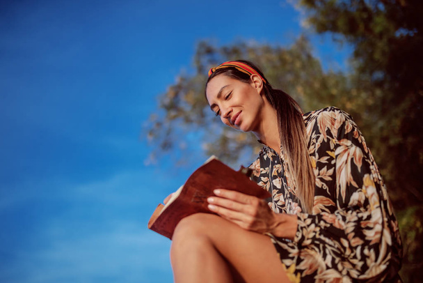 Retrato de morena caucasiana atraente e pensativa em vestido floral e com headband sentado no rock na costa ao lado do rio e escrevendo diário
. - Foto, Imagem