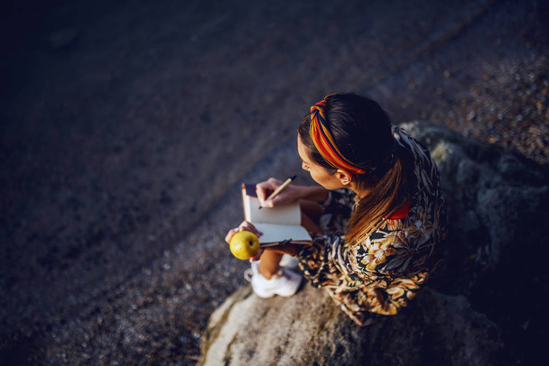 Widok z góry atrakcyjnej białej brunetki w kwiatowej sukience i z opaską siedzącą na skale na brzegu, jedzącą jabłko i piszącą pamiętnik. - Zdjęcie, obraz