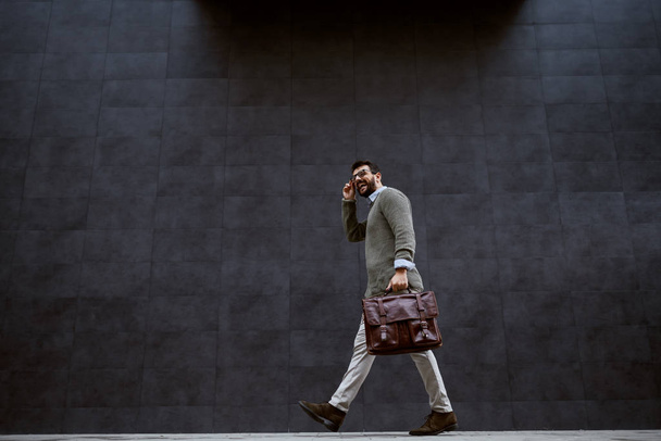 Full length of stylish caucasian designer in gray sweater holding leather bag while passing by gray wall. - Photo, Image
