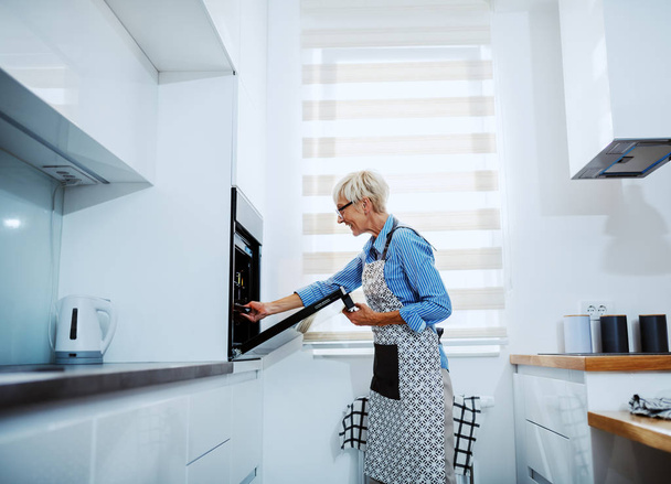 Side view of attractive caucasian senior blond woman in apron standing in kitchen and taking out baked dish from oven. - Photo, Image