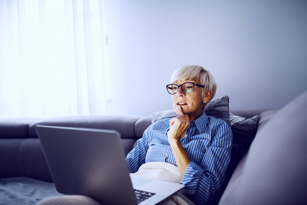 Attraente donna bionda caucasica anziana seduta sul divano coperta di coperta e guardando film sul computer portatile. Domenica pomeriggio a casa
. - Foto, immagini