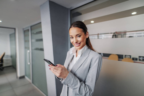 Young smiling successful gorgeous caucasian businesswoman standing in hall of corporate firm, holding smart phone and taking a break. - Фото, изображение