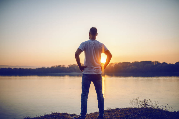 Vue arrière du bel homme caucasien debout avec les mains sur les hanches sur la falaise et regardant la rivière et le beau coucher de soleil
. - Photo, image