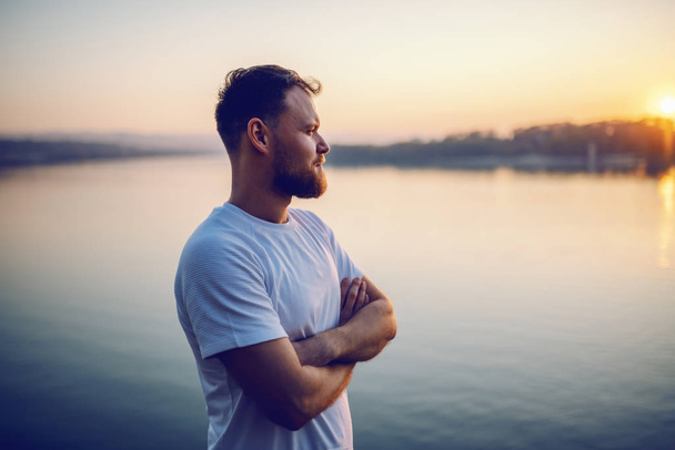 Przystojny blondyn, biały mężczyzna stojący na klifie z skrzyżowanymi rękami i patrzący na piękny zachód słońca. W tle jest rzeka. - Zdjęcie, obraz
