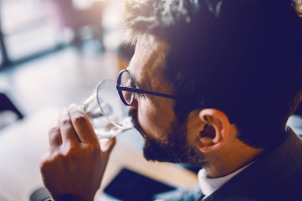 Close up of handsome thirsty caucasian bearded businessman in suit with eyeglasses sitting in cafe and drinking fresh water. - Photo, image