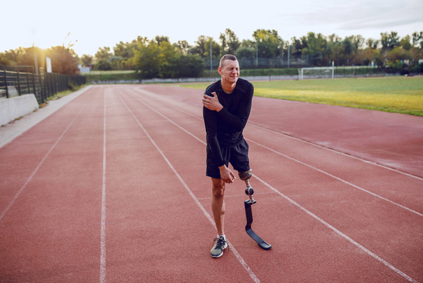 Αθλητικός καυκάσιος άνδρας με τεχνητό πόδι που στέκεται στην πίστα και έχει πόνο στο στομάχι. - Φωτογραφία, εικόνα