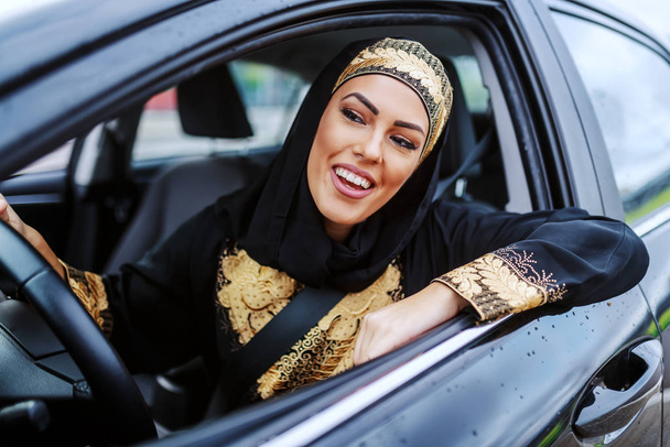Atractiva paciente sonriendo joven musulmana sentada en su coche y esperando en el atasco de tráfico
. - Foto, Imagen
