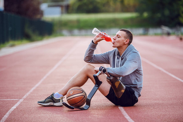schöner kaukasischer sportlich-behinderter Mann in Sportkleidung, der auf der Rennstrecke sitzt und Erfrischung trinkt. Zwischen den Beinen ist Basketballball. - Foto, Bild