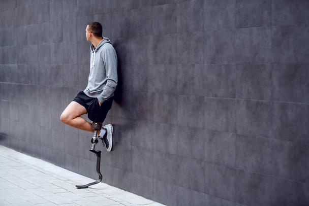 Повна довжина красивого білоруського спортсмена зі штучною ногою, що спирається на стіну зовні і має руки в кишенях
. - Фото, зображення