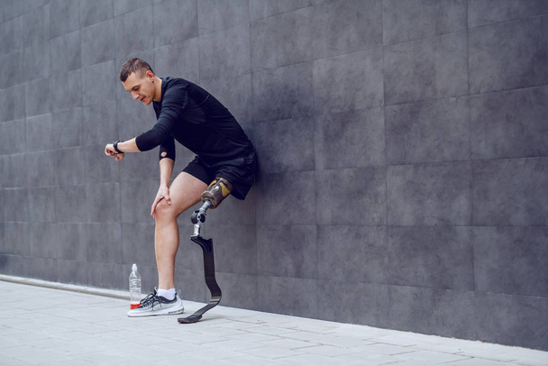 Full length of handsome caucasian sportsman with artificial leg leaning on wall, looking at wristwatch and resting from running. Next to him is bottle with refreshment. - Foto, imagen