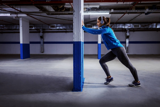 Side view of focused caucasian sportswoman in tracksuit doing pushing wall exercise in garage and listening music. Urban life concept. - Photo, Image