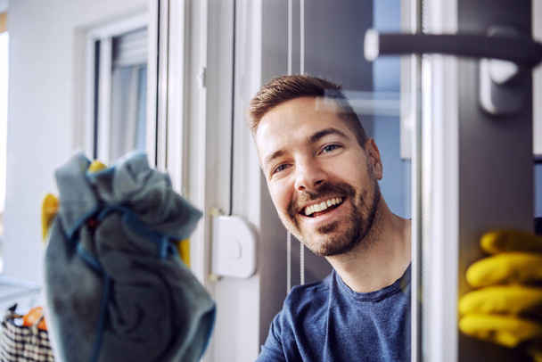 Portrait de jeune homme barbu souriant digne avec des gants en caoutchouc sur la fenêtre de nettoyage à la maison
. - Photo, image