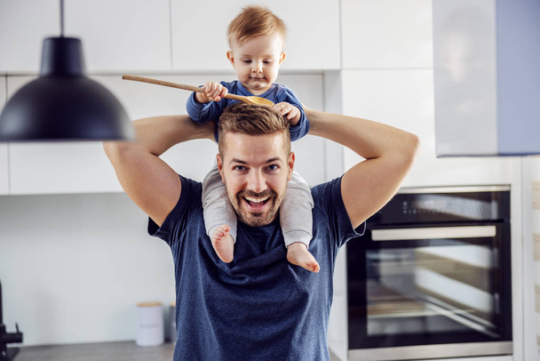 Young proud smiling positive single dad playing holding his beloved son on shoulders and having fun and kitchen. Toddler holding mixing spoon and playing with it. - Foto, Imagen