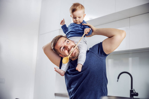 Young handsome single dad holding his only son on his shoulder, looking at him and biting mixing spoon. Toddler holding mixing spoon and looking at dad. - Valokuva, kuva