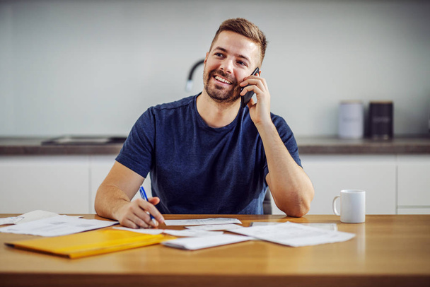 Jonge aantrekkelijke knappe bebaarde glimlachende man bellen klantenservice tijdens het zitten aan tafel en het invullen van rekeningen. - Foto, afbeelding