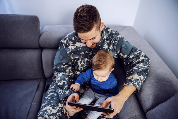 Άποψη γενναίου στρατιώτη που αναπληρώνει το χαμένο χρόνο με το γιο του. Άντρας κρατώντας το παιδί στην αγκαλιά, δισκίο στα χέρια και βλέποντας κινούμενα σχέδια. - Φωτογραφία, εικόνα