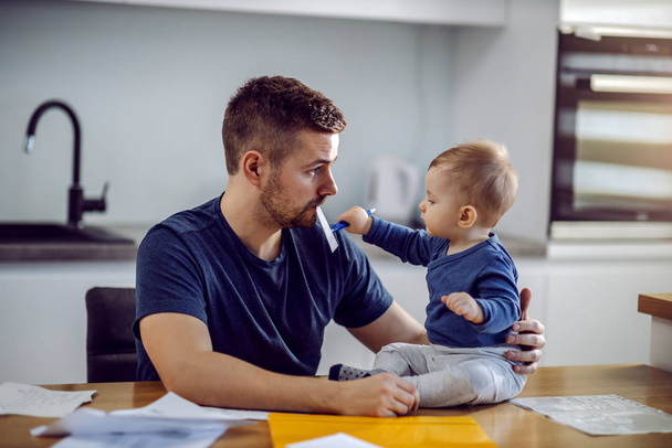 Young proud dad sitting at dining table, having bill in mouth and holding his son. Toddler is sitting on table and helping his dad to fill in bill. - Фото, изображение