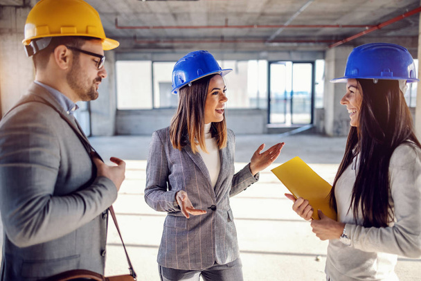 Group of young highly motivates attractive caucasian architects with helmets on heads standing in building in construction process and having casual talk on pause. - Photo, Image