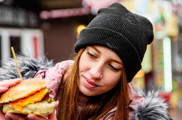 Street food. young woman holding juicy burger and eating oudoor winter - Foto, afbeelding