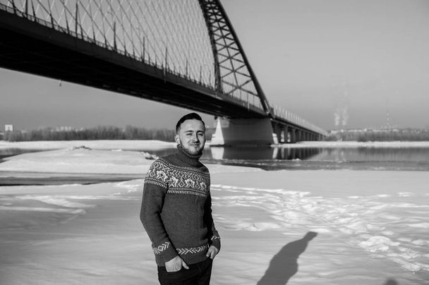 man in warm clothes with a beard is walking on the street in the winter in a warm sunny day at the river bridge in the background - Photo, Image