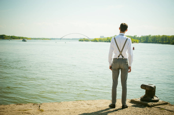 young skinny man, elegantly stylishly dressed in white shirt, gray trousers with suspenders and bow tie. Looking away, at the river water bridge and ship - Zdjęcie, obraz