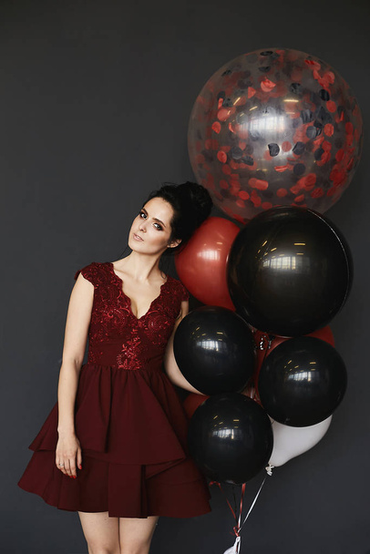 Young happy brunette woman in modish red dress with big balloons in her hand posing at grey background, isolated. Sexy model girl with perfect body posing with colorful balloons for Valentines day - Φωτογραφία, εικόνα