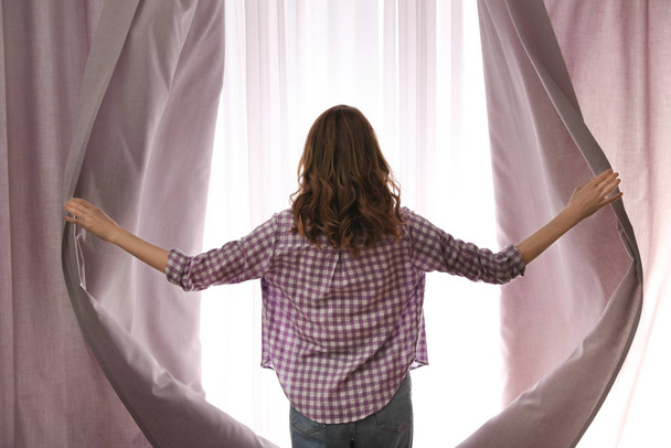 Woman opening window curtains at home in morning, back view - Photo, image