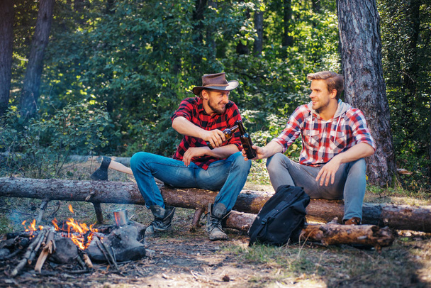 Two handsome men friends on camping near campfire. Tourists relaxing. Two friends lumberjack worker sitting in the forest with beer. Friends enjoy weekend camp in forest. - Foto, Bild