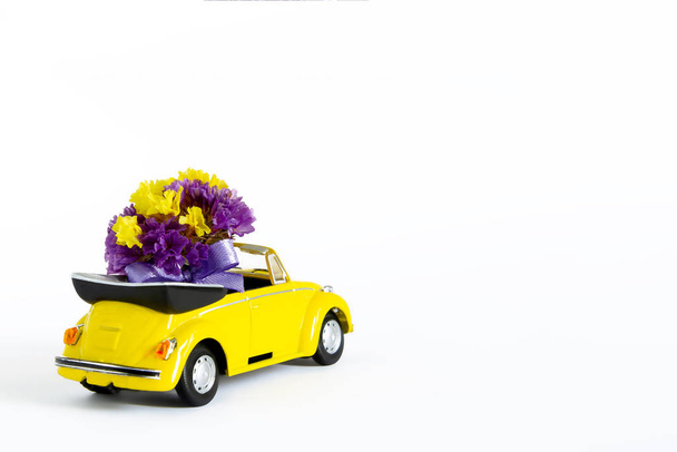 View of a colorful bouquet of purple flowers that is in a small yellow retro car on a white background. Selective focus. The concept of a holiday, wedding, flower delivery, gift - Fotografie, Obrázek