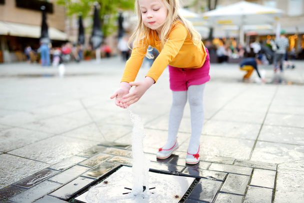 Sıcak ve güneşli yaz gününde şehir çeşmesinin yanında oynayan tatlı küçük kız. Çocuk yazın suyla eğleniyor.. - Fotoğraf, Görsel